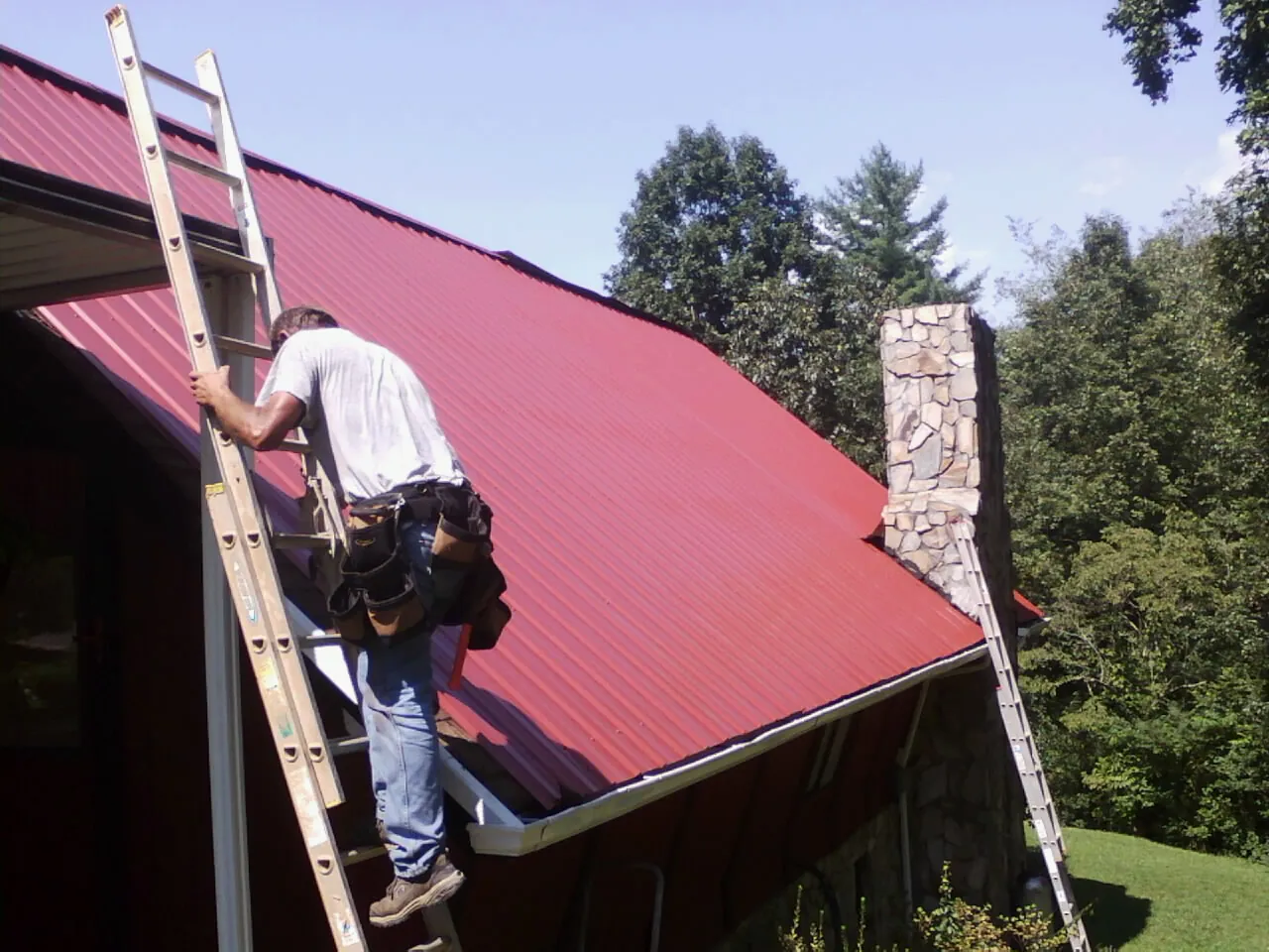 Roofer on a ladder beside a red metal roof and stone chimney — Soffit & Fascia Repair by Roofing Brothers in Folcroft